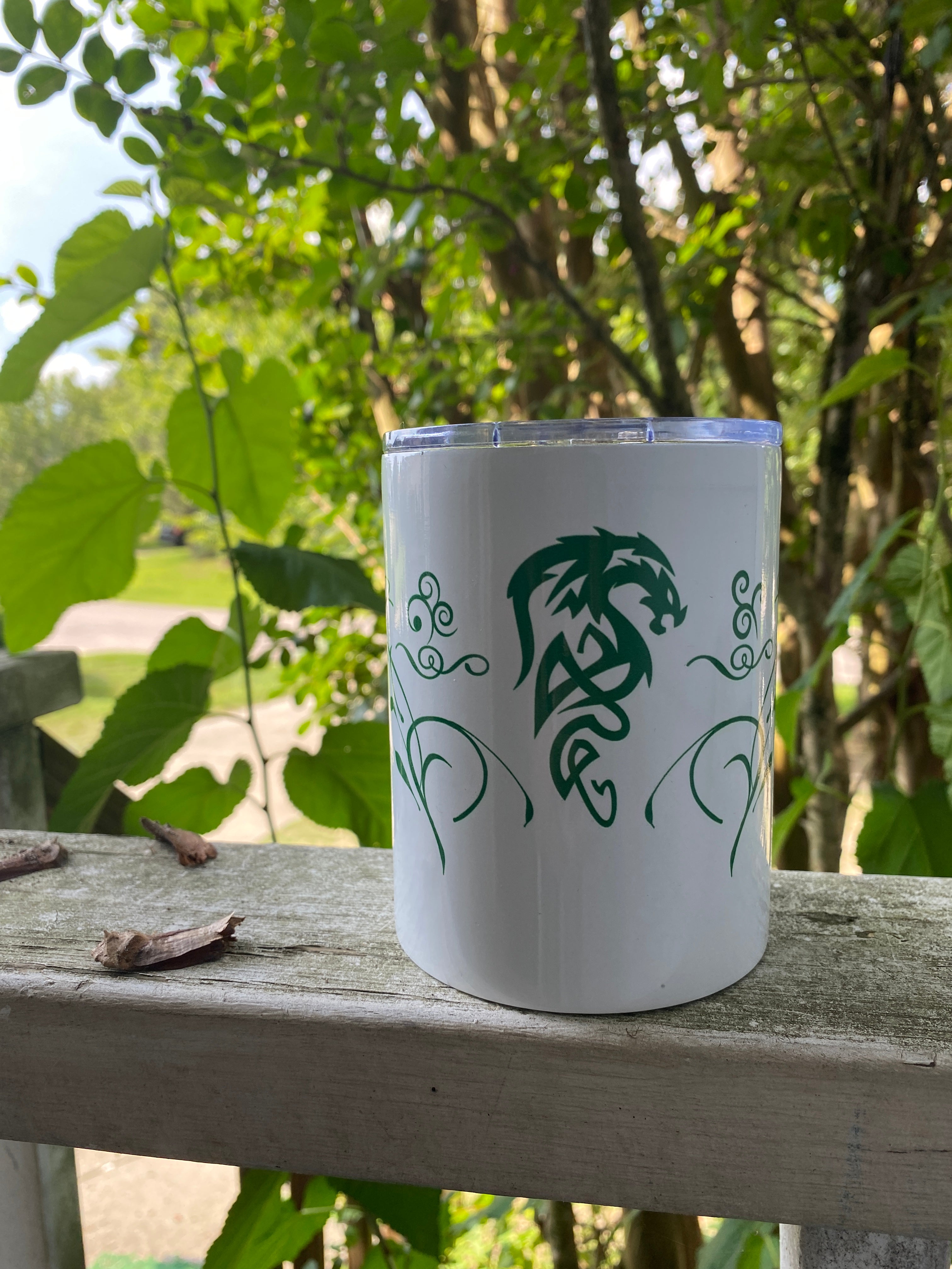The 10oz tumbler sitting outside on a ledge with beautiful, lush greenery behind it. It has a dragon design