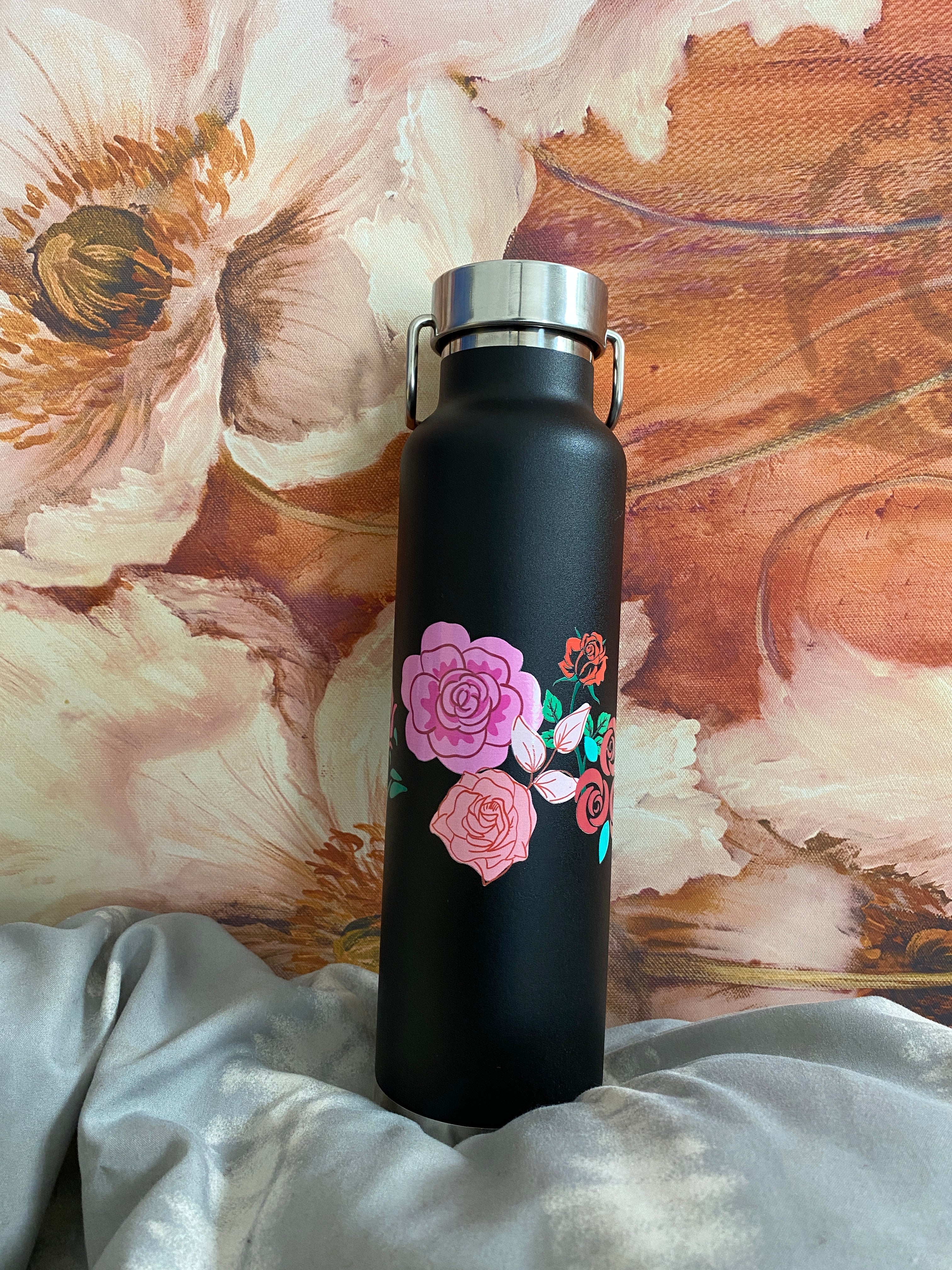 The 22oz copper vacuum insulated bottle in the black color with a flowers and rose design. It is positioned in front of artwork that also has flowers on it.